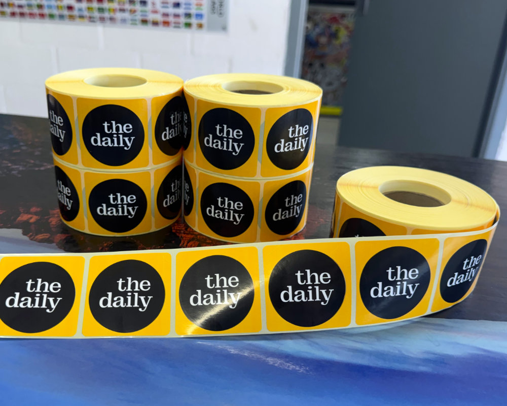 Rolls of glossy yellow "the daily" circular labels stacked and unrolled on a table